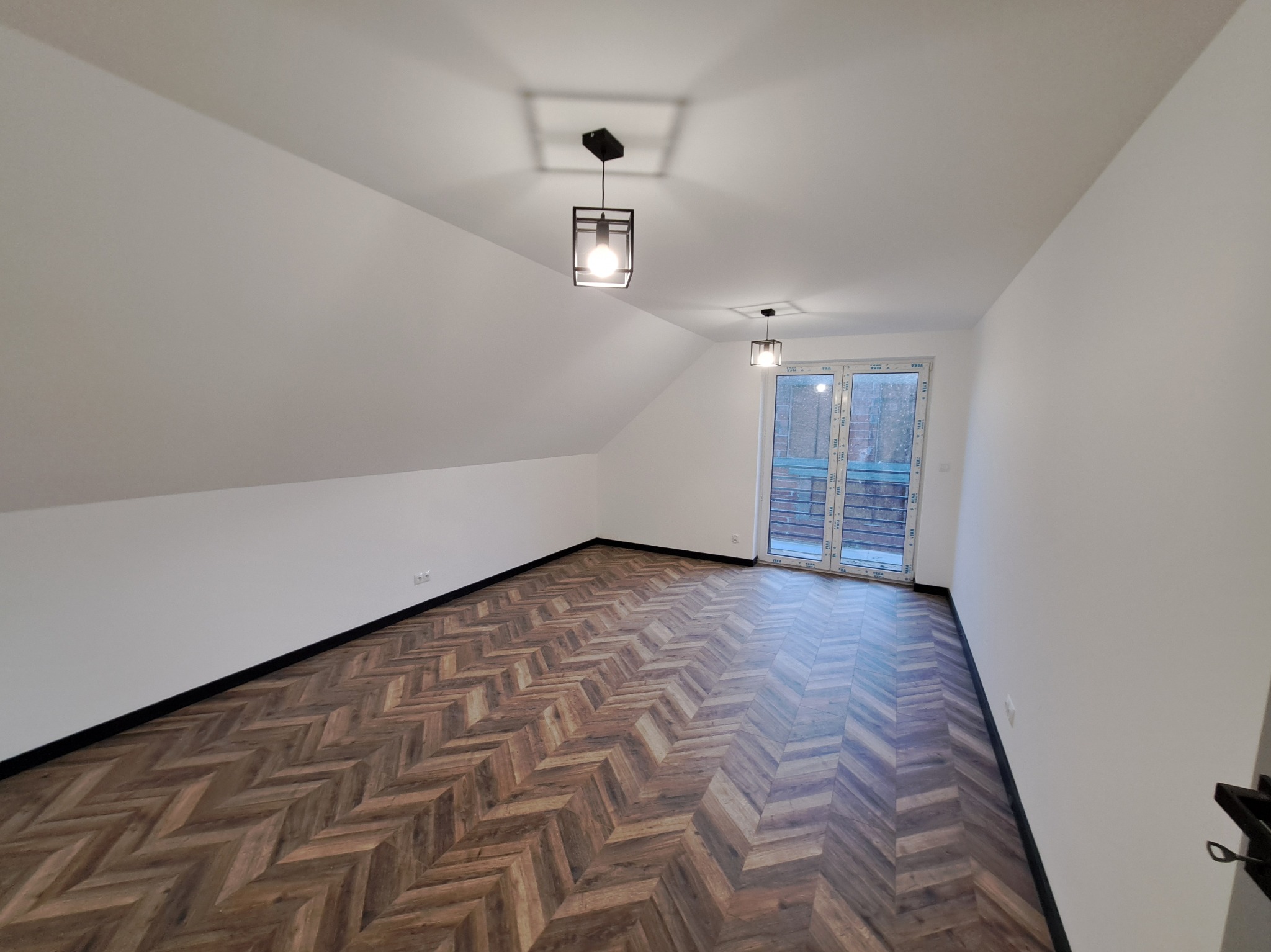 Poddasze z parkietem w jodełkę, białe ściany i czarne listwy przypodłogowe. Dwa nowoczesne, czarne plafony. Okno balkonowe z widokiem na zieleń.