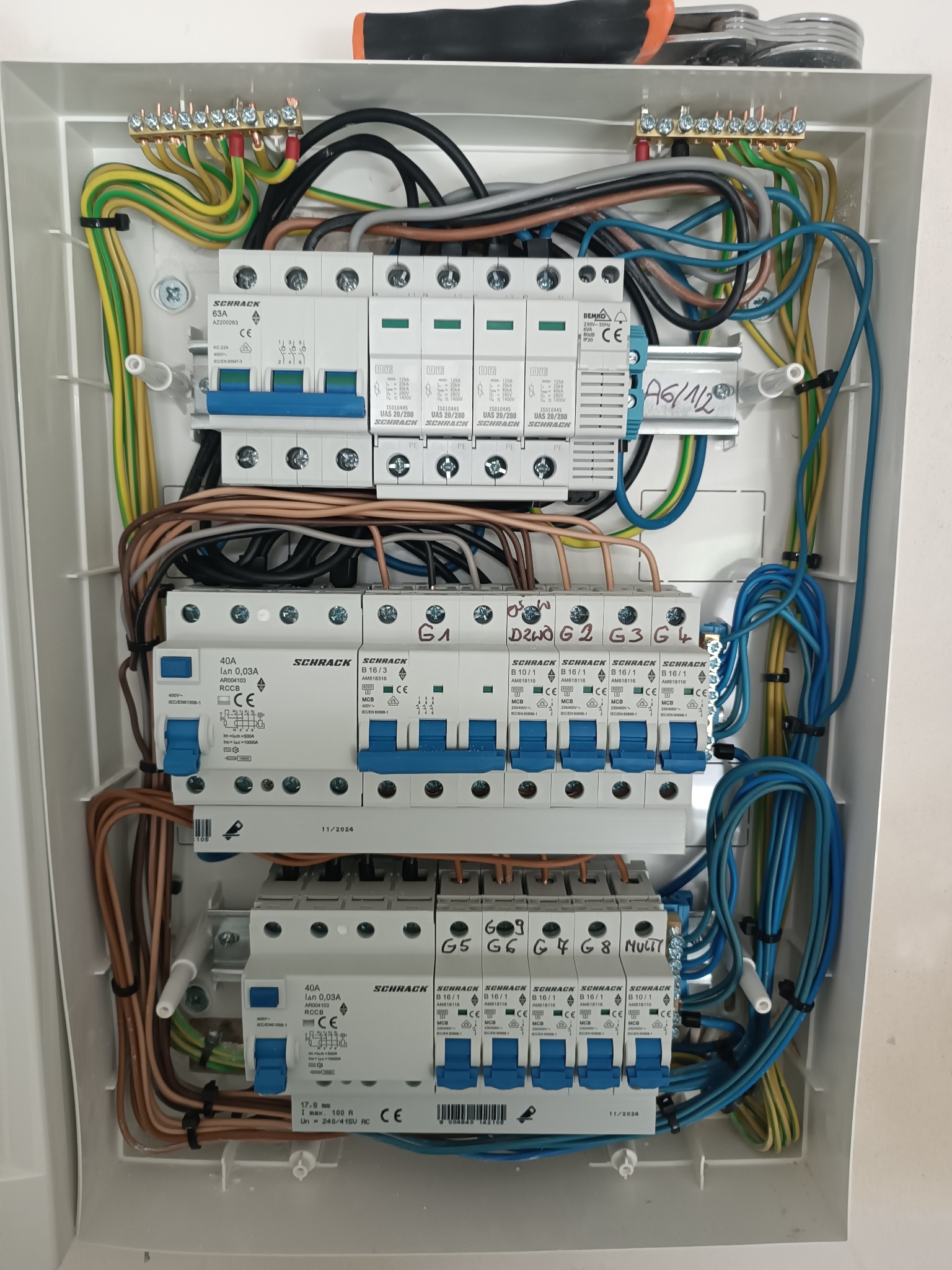 Rozdzielnica elektryczna Schrack z zainstalowanymi bezpiecznikami i okablowaniem, widoczna z góry, z narzędziem w tle. Element instalacji elektrycznej wewnątrz obudowy.