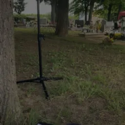 Nagłośnienie na cmentarzu z mikrofonem i tekstem piosenki, prawdopodobnie podczas uroczystości pogrzebowej. Sprzęt ustawiony obok drzewa, w tle nagrobki.
