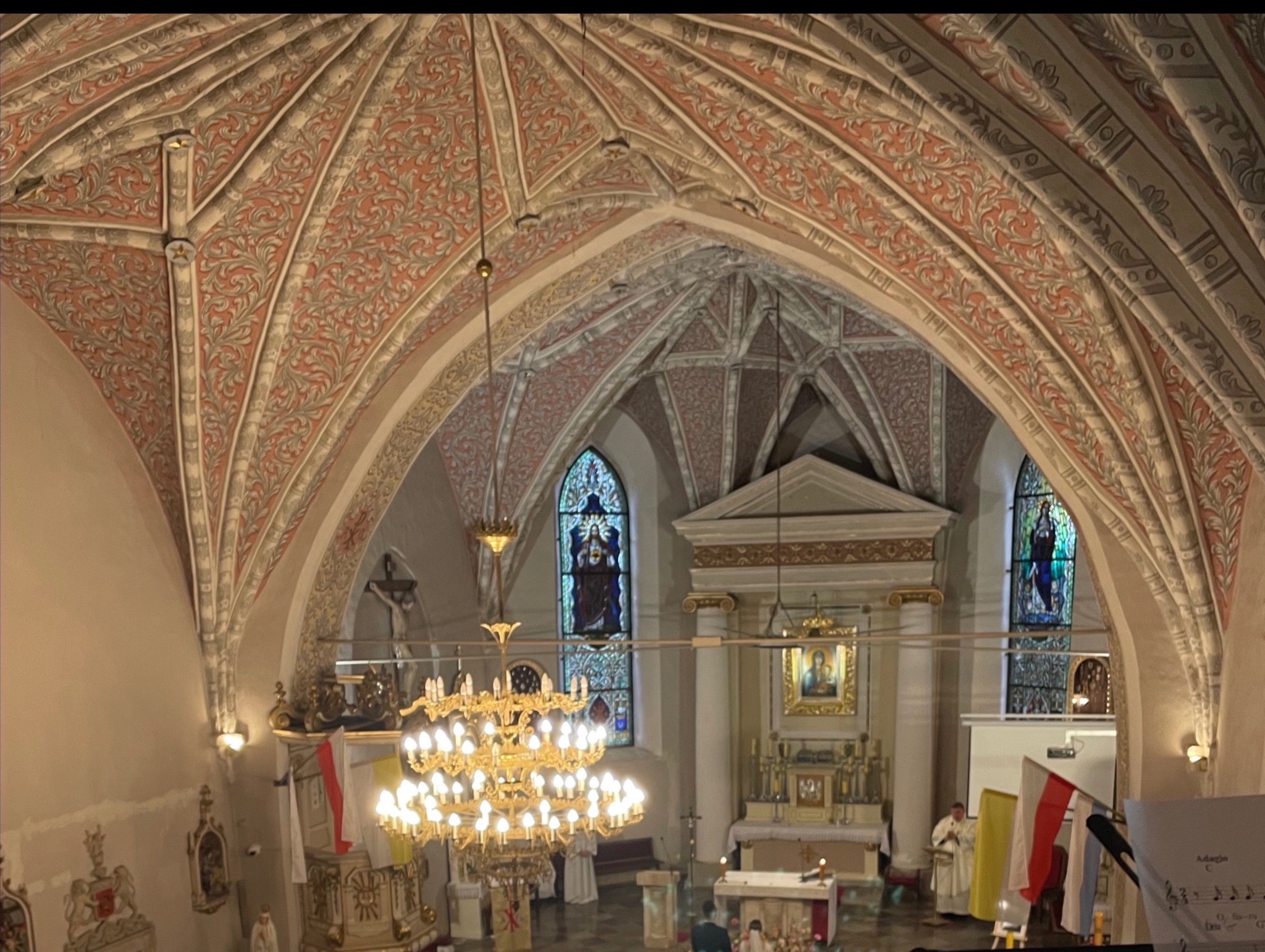 Wnętrze kościoła z bogato zdobionym sklepieniem, witrażami i żyrandolem. Widoczny ołtarz i osoby podczas uroczystości. Flagi Polski i Watykanu.