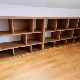 Lignum Art MP corp sp. z o.o. - Długa, drewniana szafa na wymiar z licznymi przegródkami, wykonana z jasnego drewna, ustawiona w pomieszczeniu z parkietem. Widok z dołu, podkreślający przestronność.