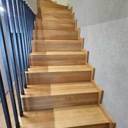 Lignum Art MP corp sp. z o.o. - Jasne, drewniane schody z czarną, metalową balustradą, prowadzące w górę. Minimalistyczny design, szara ściana w tle. Widok z dołu, podkreślający geometrię stopni.