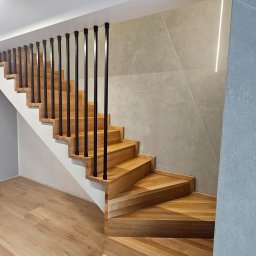 Lignum Art MP corp sp. z o.o. - Kątowe ujęcie drewnianych schodów z czarną balustradą, oświetlonych listwą LED na ścianie. Parkiet w odcieniach beżu i szarości. Nowoczesny design.