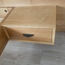 TREZZA - Jasna, drewniana szafka nocna z szufladą i półokrągłym uchwytem, widoczna na tle drewnianej ściany i podłogi w odcieniach szarości. Realizacja z Krakowa.