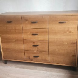 TREZZA - Drewniana komoda z szufladami i szafkami, z ciemnymi uchwytami, stojąca w pokoju obok drzwi i rośliny doniczkowej. Jasne drewno, minimalistyczny design.