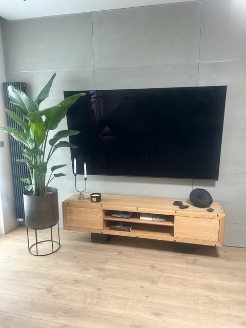 Jasna, drewniana szafka RTV z półkami, na tle szarej ściany z telewizorem. Obok stoi roślina w doniczce. Minimalistyczny design wnętrza z drewnianą podłogą.