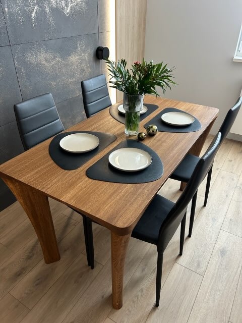 Drewniany stół z czarnymi krzesłami i dekoracjami na blacie, na tle ściany z efektem betonu i drewnianej lameli z oświetleniem. Nowoczesne wykończenie wnętrza.