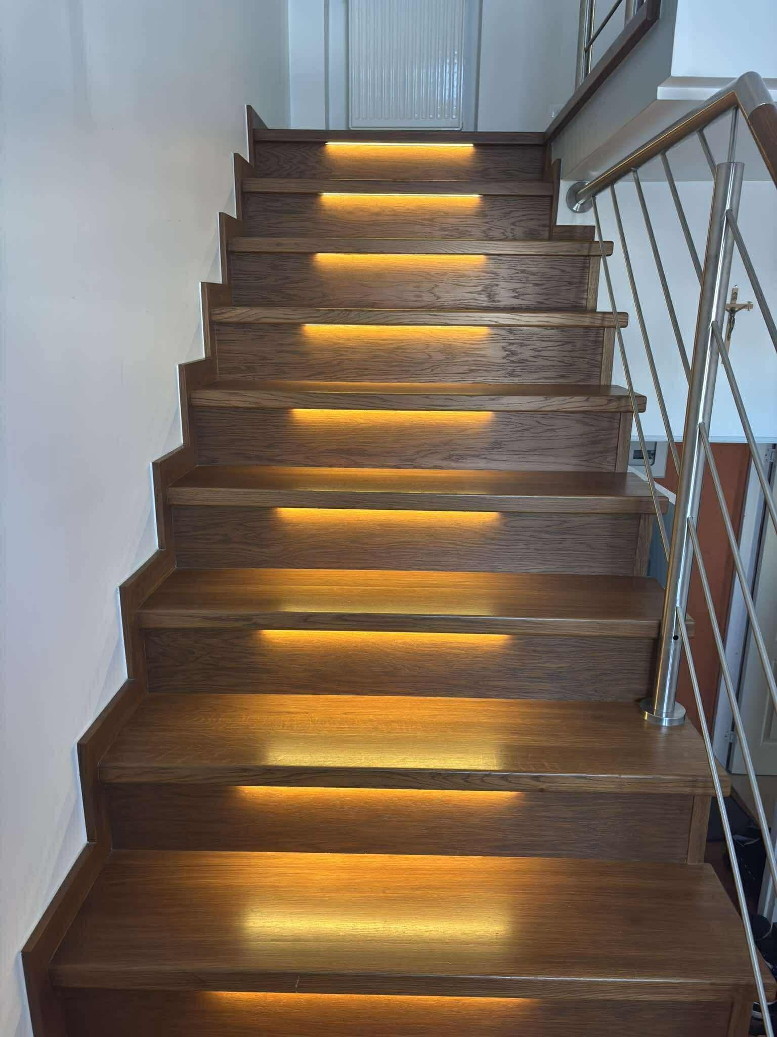 Drewniane schody z podświetleniem LED na każdym stopniu, z metalową balustradą. Ujęcie od dołu, podkreślające nowoczesny design i ciepłe światło.