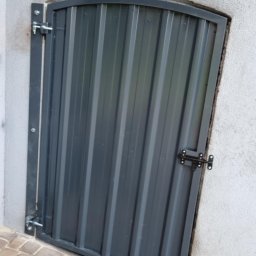 TobiWelding - Szara, metalowa furtka z zamkiem i pionowymi panelami, wbudowana w białą ścianę, z kostką brukową u dołu. Furtka ma zaokrąglone górne rogi.