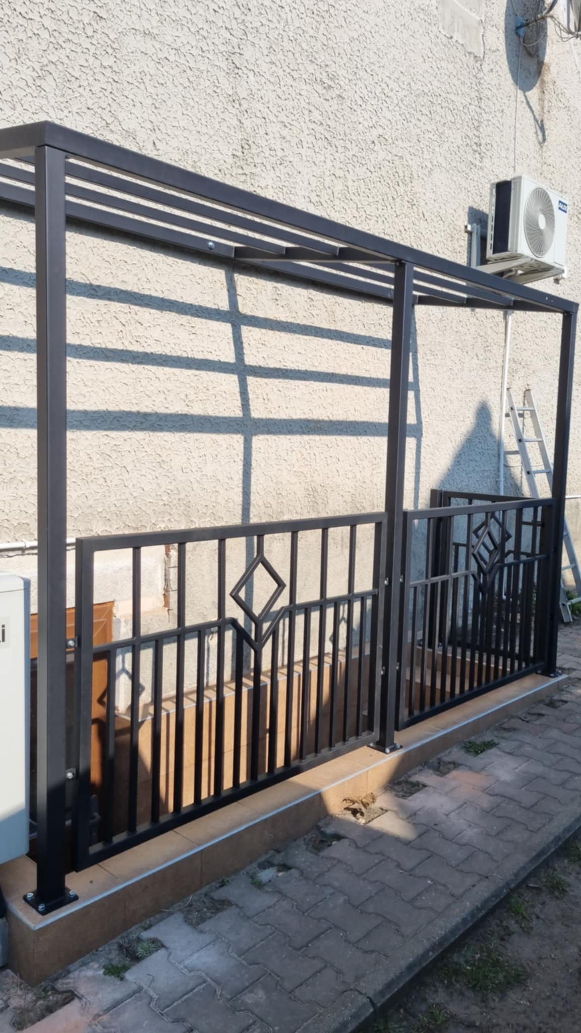 Ciemna metalowa pergola z dekoracyjnymi elementami ogrodzenia, przylegająca do ściany budynku. Widoczne cienie rzucane przez konstrukcję i drabina oparta o ścianę.