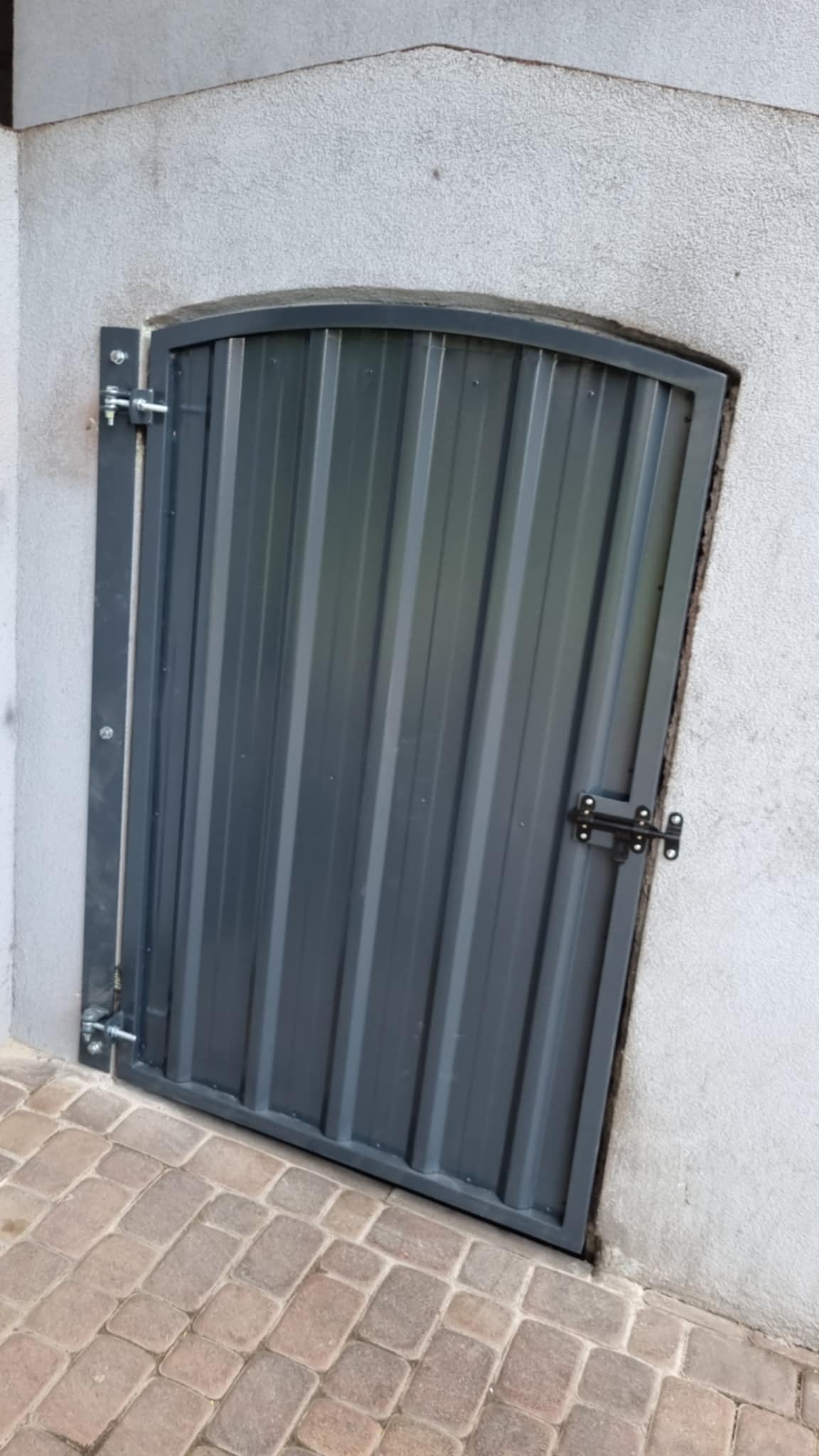 Szara, metalowa furtka z zamkiem i pionowymi panelami, wbudowana w białą ścianę, z kostką brukową u dołu. Furtka ma zaokrąglone górne rogi.
