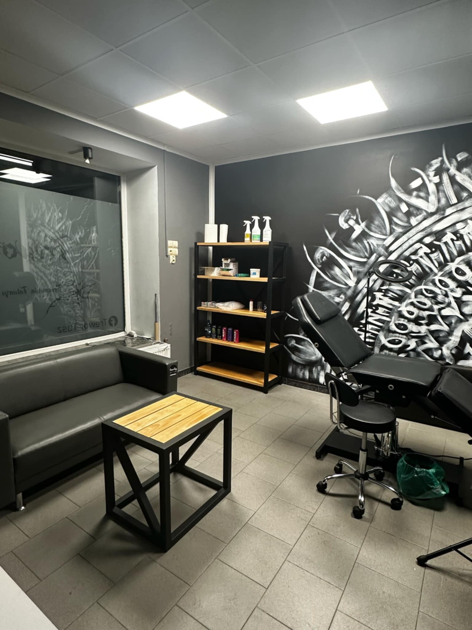 Nowoczesne studio tatuażu: czarny fotel, grafika na ścianie, drewniany stolik. Minimalistyczny design z industrialnymi akcentami. Widok na czyste, dobrze oświetlone wnętrze.