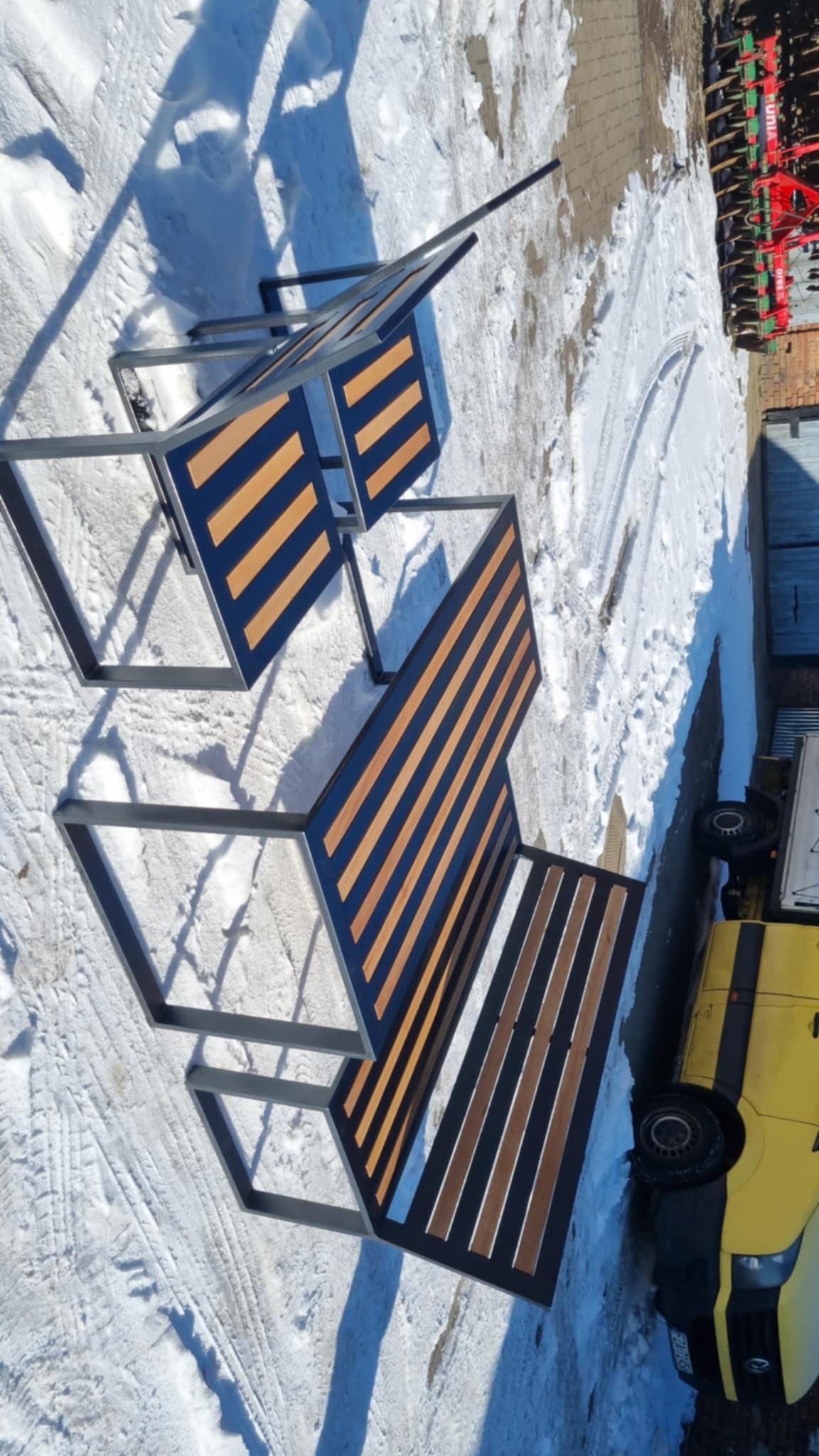 Nowoczesna pergola metalowa z drewnianymi listwami w kolorze brązowym, ustawiona na śniegu. Widoczny fragment żółtego samochodu dostawczego i maszyna rolnicza w tle.