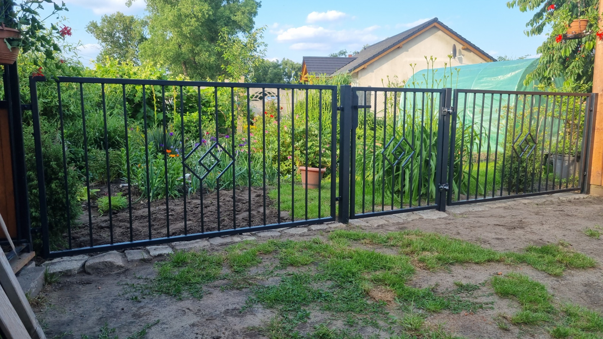 Czarna metalowa brama wjazdowa z dekoracyjnymi rombami, prowadząca do bujnego ogrodu z kwiatami i zielenią, na tle domu z ogrodem.