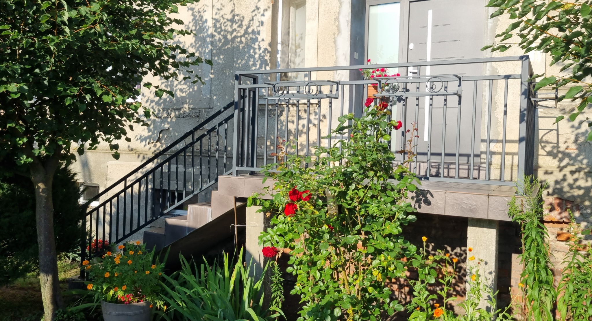 Metalowa balustrada schodowa i balkonowa w szarym kolorze, z dekoracyjnymi elementami, otoczona zielenią i kwiatami. Widok na wejście do domu.