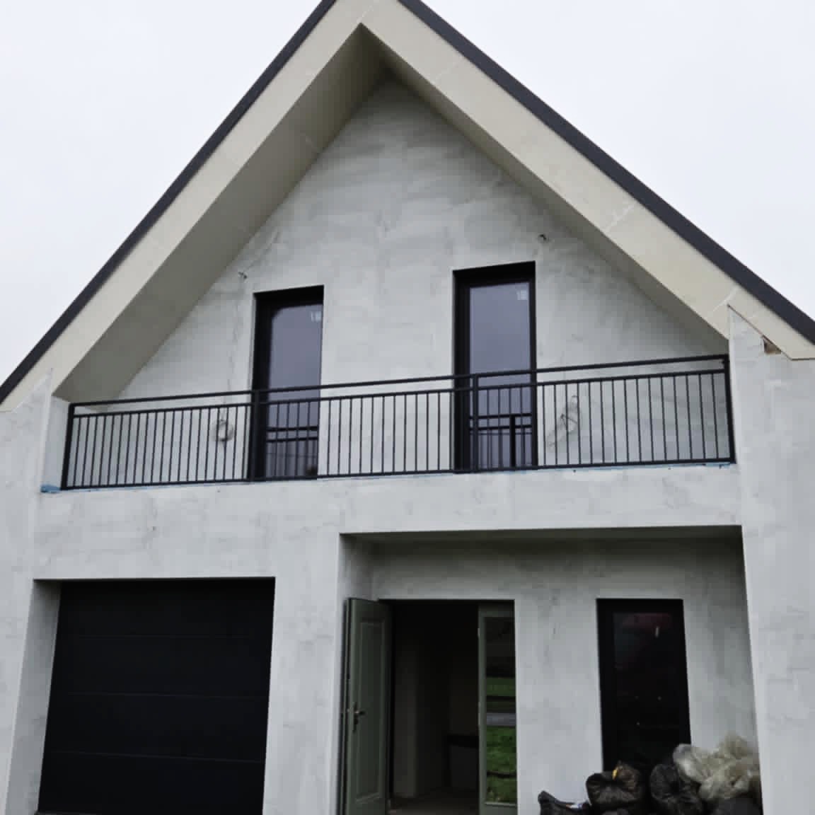 Nowoczesny dom z balkonem zabezpieczonym czarną, metalową balustradą. Minimalistyczny design, surowy beton elewacyjny. Widok z dołu na dwukondygnacyjny budynek.