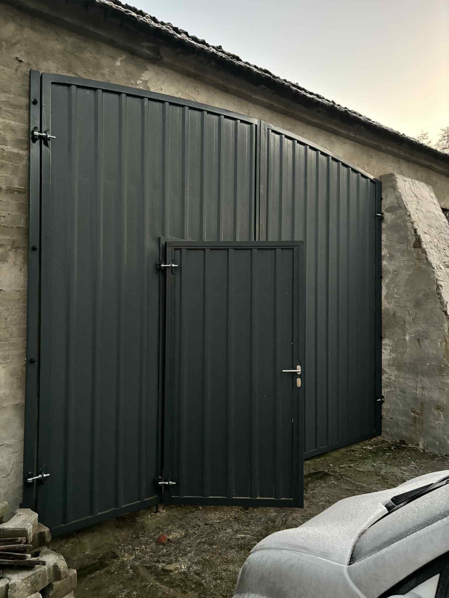 Nowoczesna, grafitowa brama wjazdowa z furtką, wykonana z blachy trapezowej. Widoczne detale mocowań i zamek. Tło stanowi fragment budynku z surowego betonu.