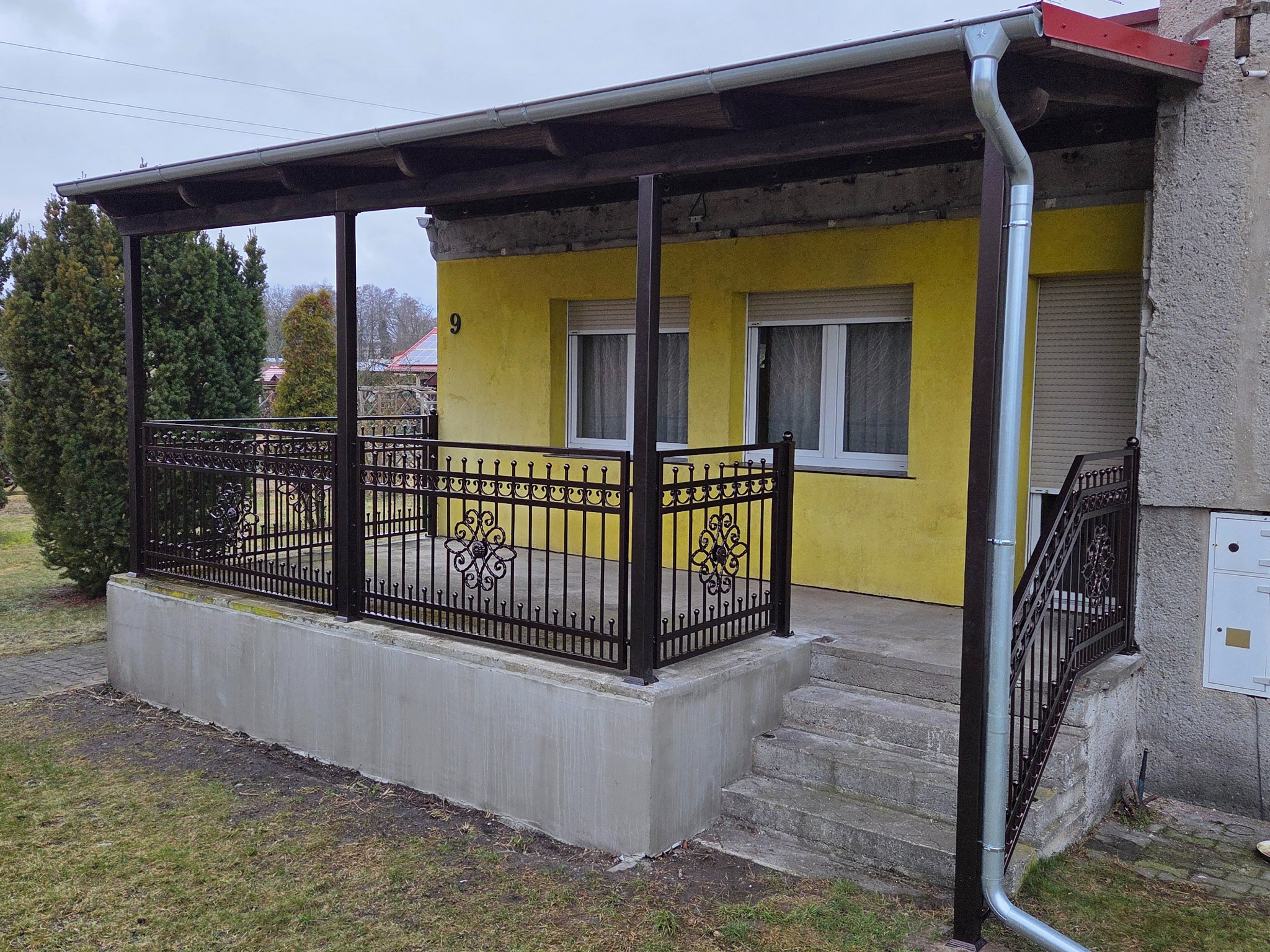Zadaszony taras z kutą, czarną balustradą o dekoracyjnych motywach kwiatowych, na tle żółtej elewacji budynku z widocznym numerem 9 i fragmentem rynny.