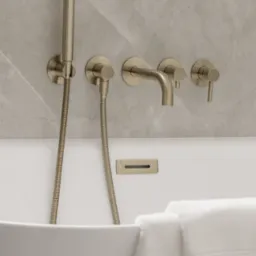 Bateria wannowa w kolorze szczotkowanego złota, zamontowana na marmurowej ścianie nad białą wanną z ułożonymi ręcznikami. Minimalistyczny design i eleganckie wykończenie.