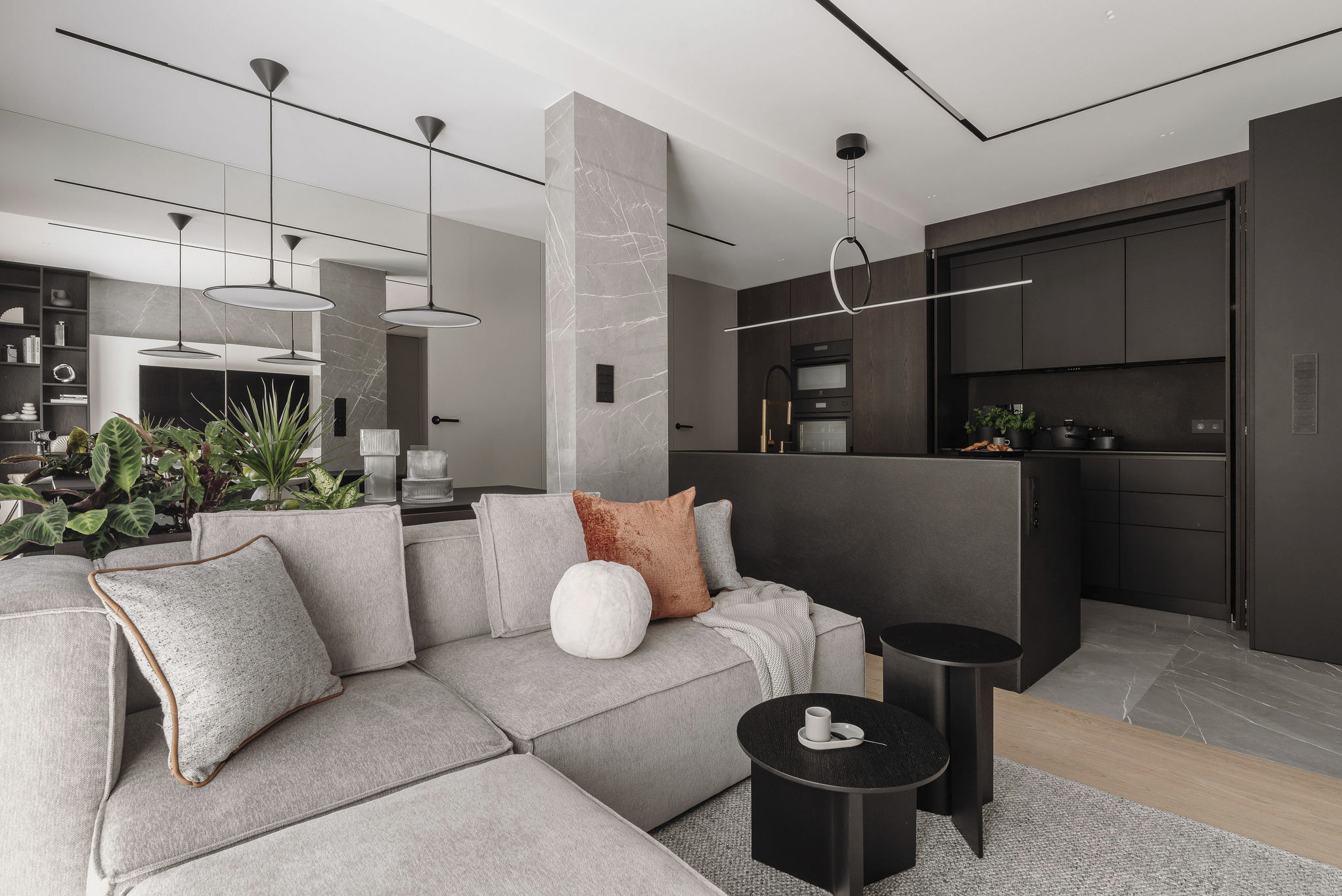 Nowoczesne wnętrze: szary narożnik z poduszkami, stoliki kawowe, czarna kuchnia w tle, marmurowy filar i designerskie lampy wiszące. Minimalizm i elegancja.