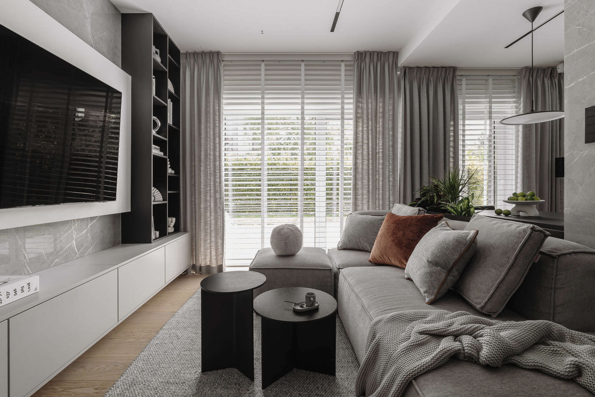 Nowoczesny salon z szarą sofą, stolikami kawowymi i telewizorem na ścianie z marmurowym wzorem. Minimalistyczne oświetlenie i zasłony w neutralnych barwach.
