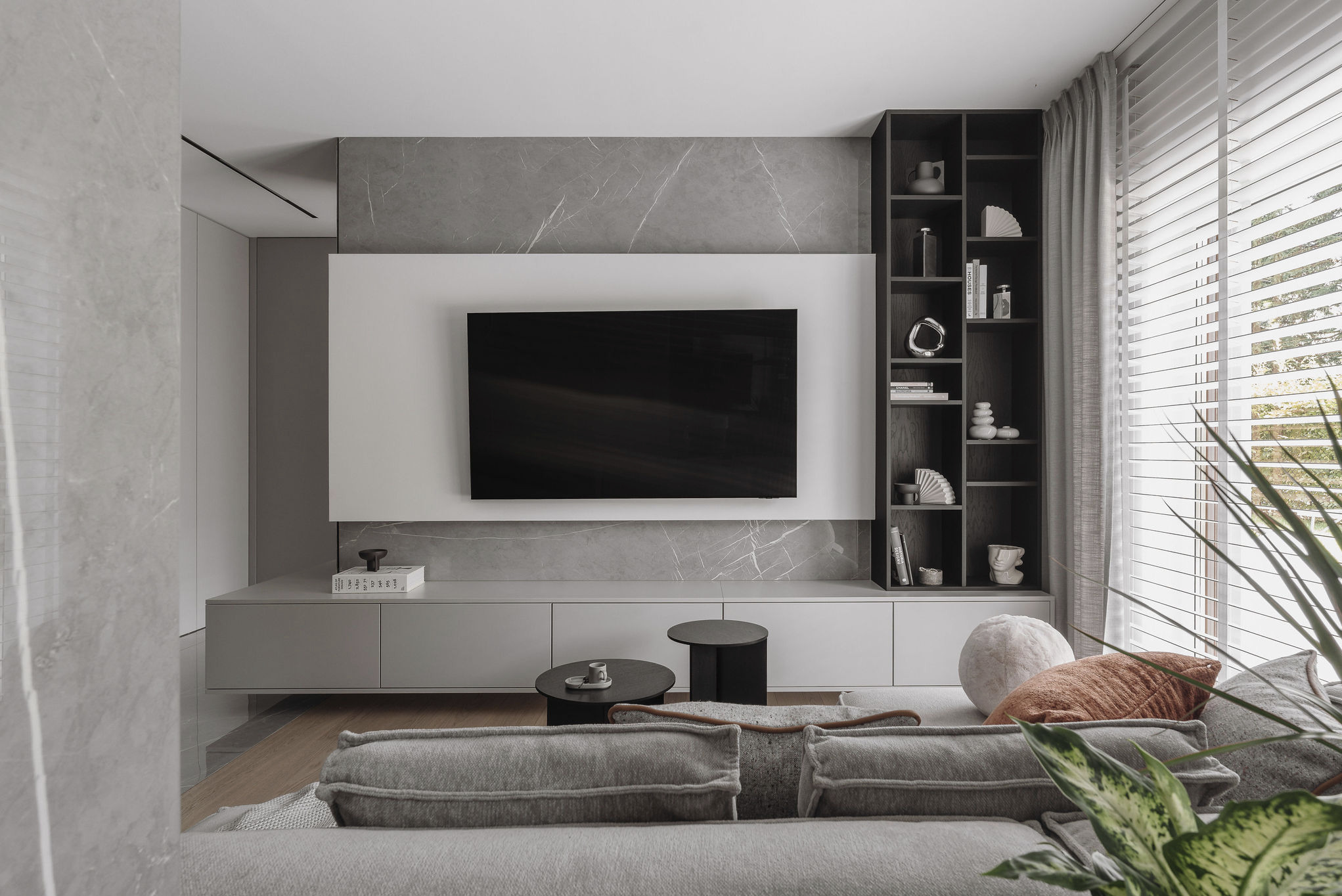 Stylowy salon z szarą kanapą, telewizorem na tle marmurowej ściany i półką z dekoracjami. Minimalistyczny design z akcentami roślinnymi i naturalnym światłem.