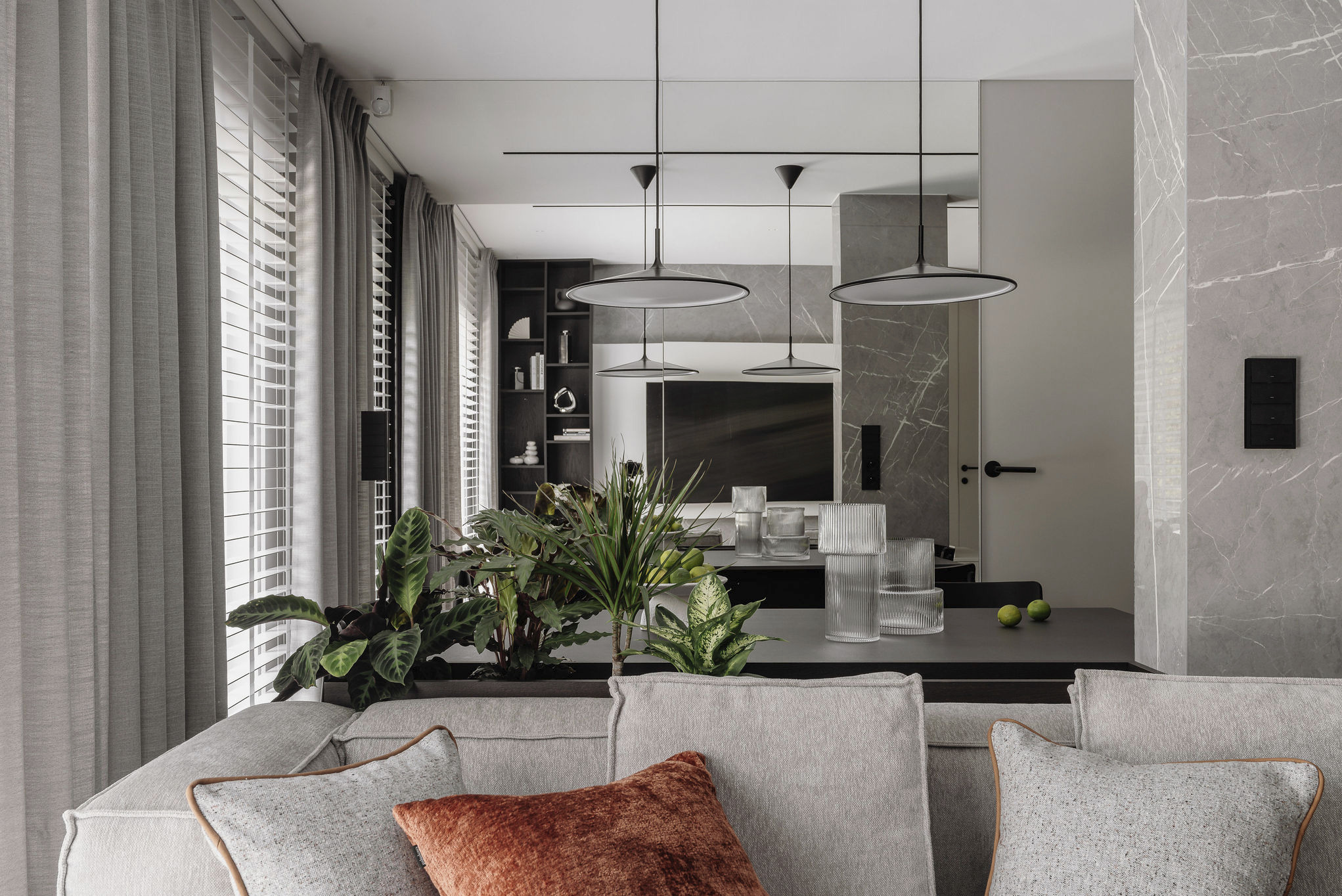 Elegancki salon z szarą sofą, roślinami doniczkowymi i minimalistycznymi lampami wiszącymi. W tle marmurowa ściana i lustro powiększające przestrzeń.