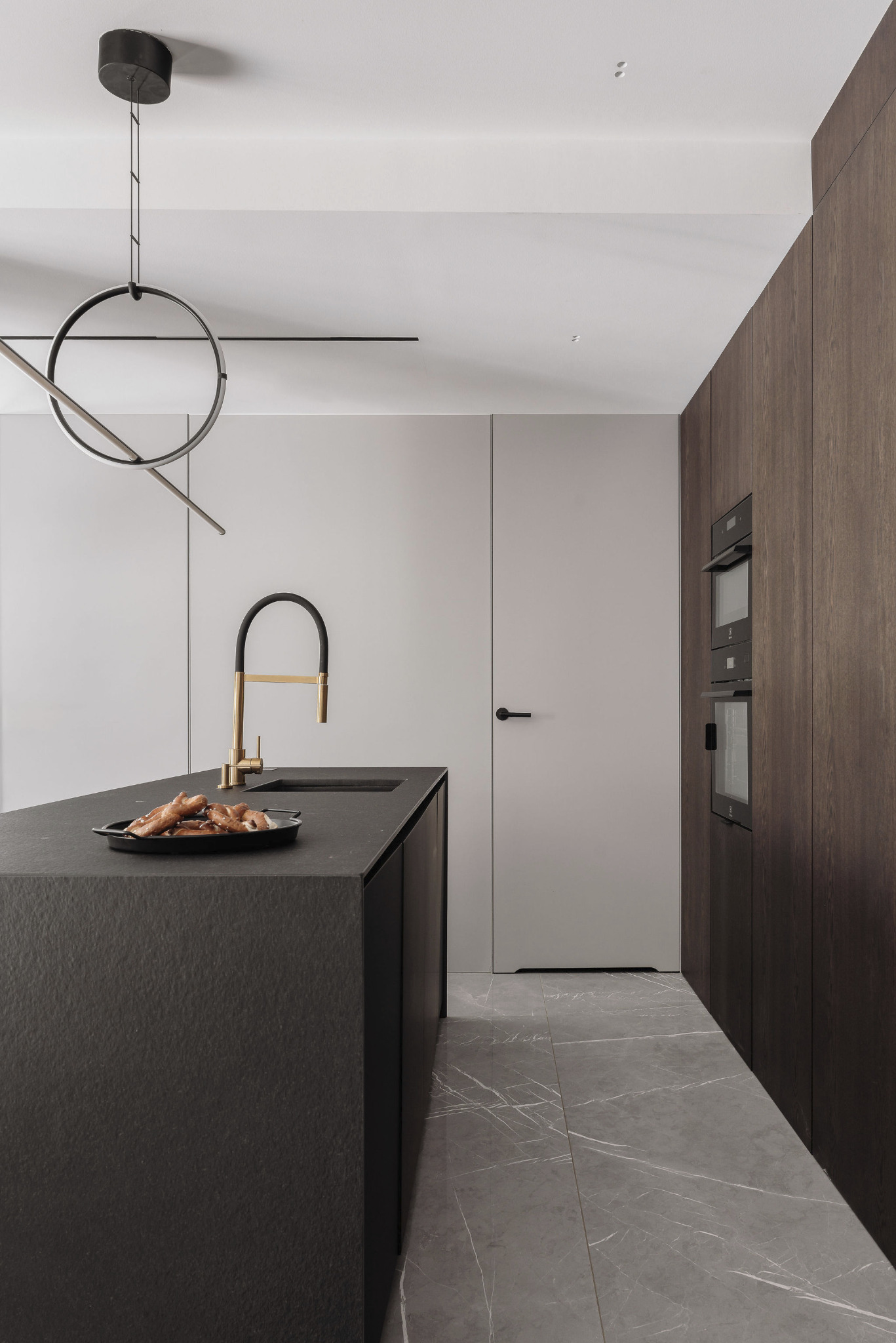 Minimalistyczna kuchnia: czarny blat wyspy z przekąskami, złota bateria, szare płytki imitujące marmur i drewniana zabudowa z piekarnikami.