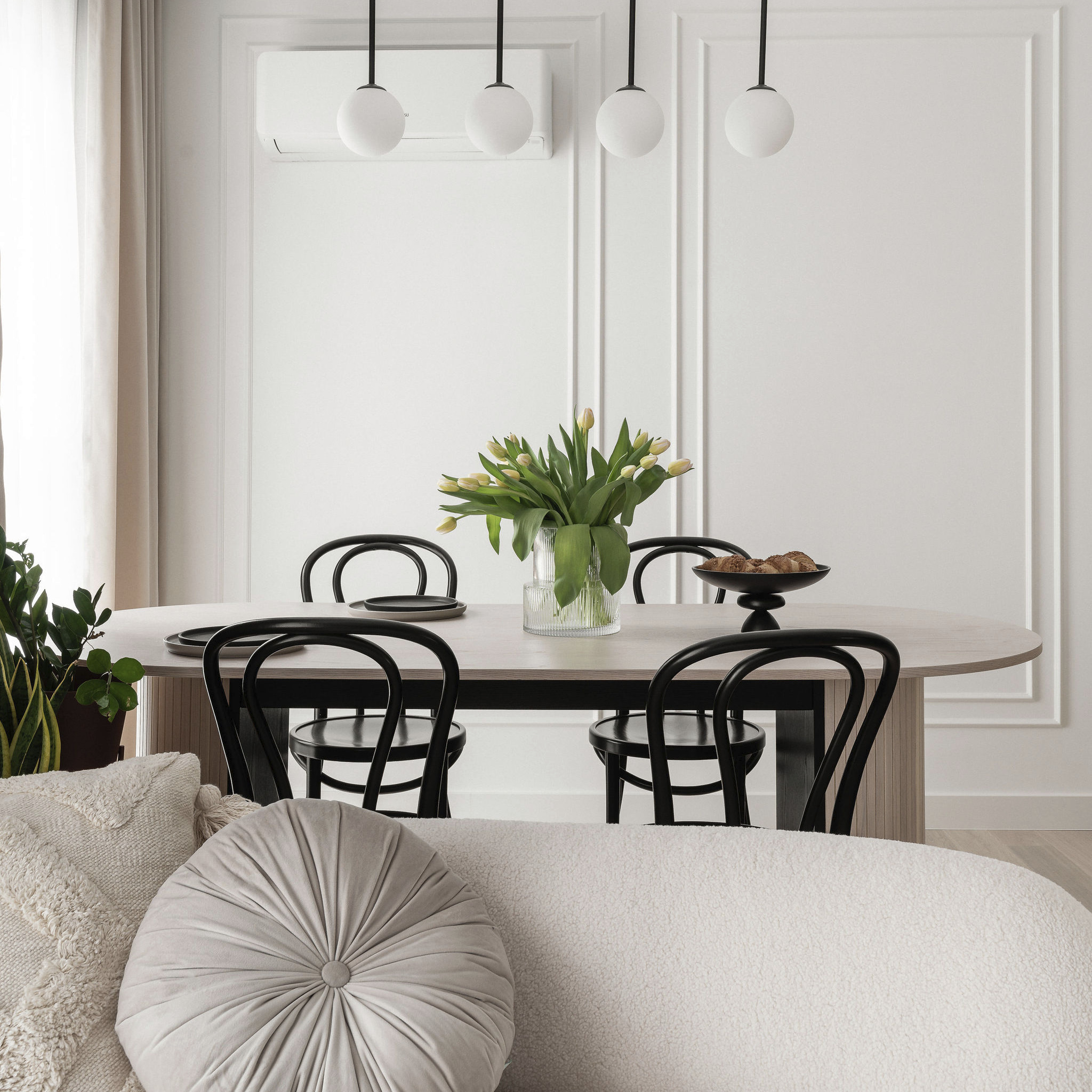 Jasne wnętrze jadalni z owalnym stołem, czarnymi krzesłami, dekoracyjnymi lampami i klimatyzatorem. Na stole wazon z tulipanami i misa z pieczywem.