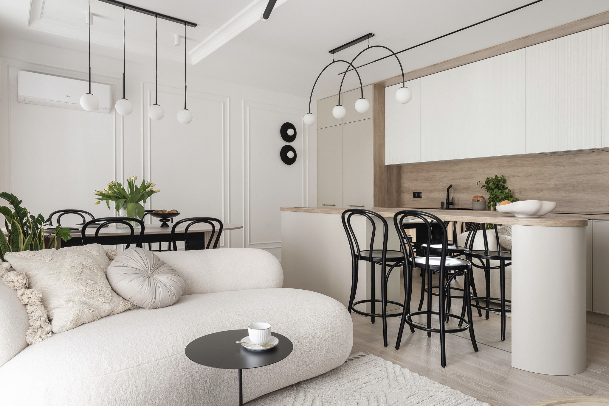 Jasne, przestronne wnętrze z aneksem kuchennym i sofą boucle. Nad wyspą kuchenną lampy z kulistymi kloszami. Minimalistyczny design z drewnianymi akcentami.