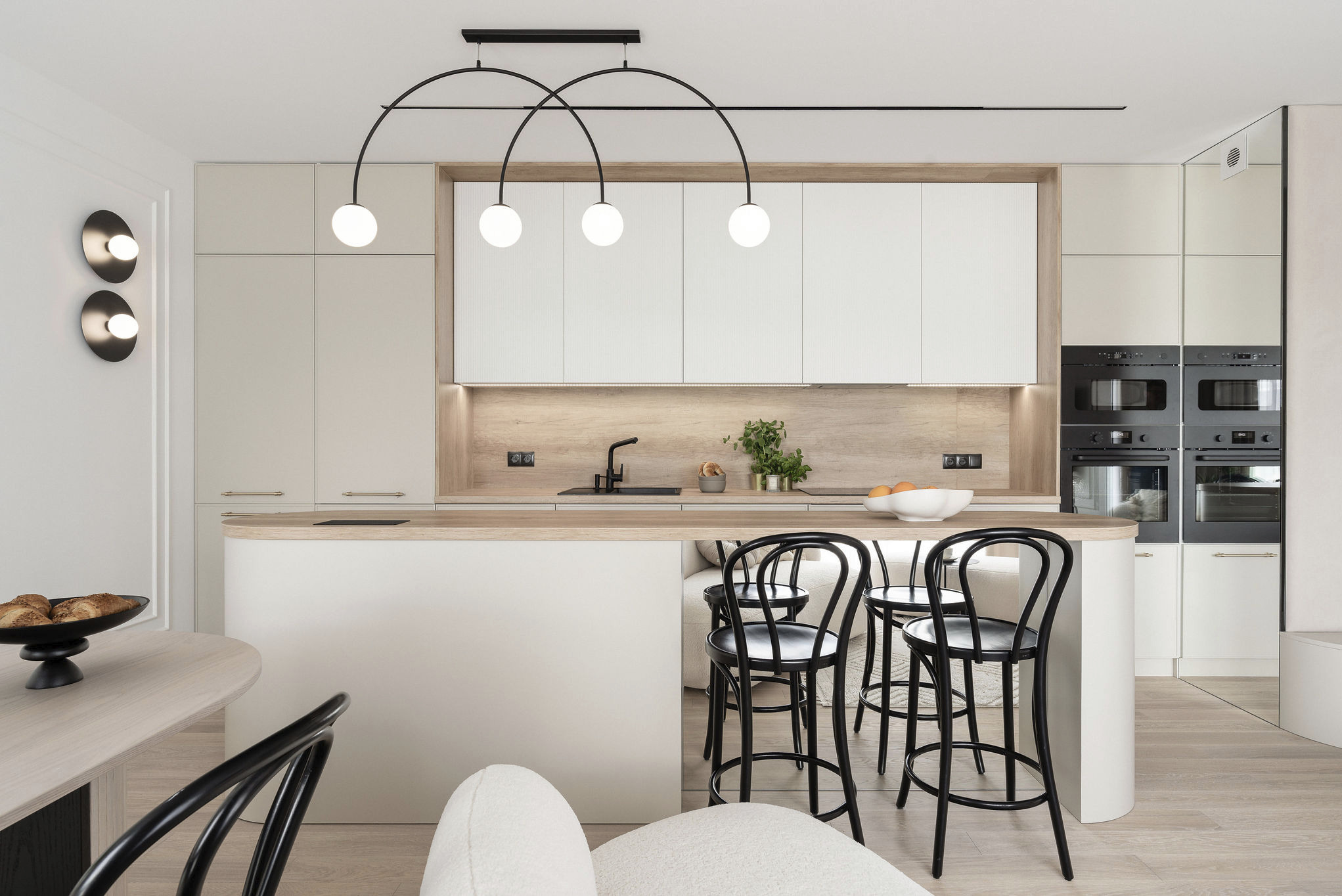 Jasna kuchnia z wyspą i czarnymi hokerami. Nad wyspą lampa z trzema kulistymi kloszami. Zabudowa kuchenna w odcieniach beżu i bieli. Dwie czarne wbudowane kuchenki.