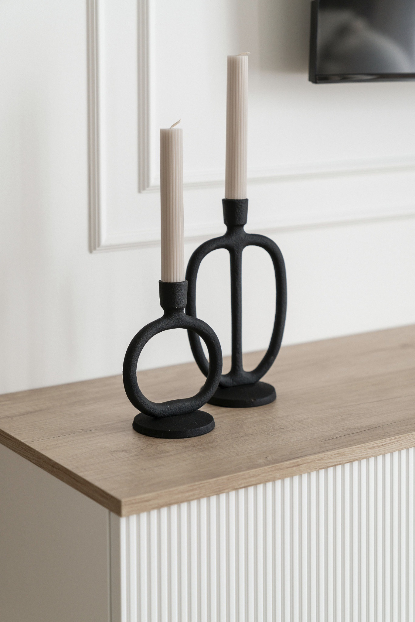 Dwa czarne, nowoczesne świeczniki z beżowymi świecami na drewnianym blacie komody z ryflowanym frontem i białym panelem ściennym.
