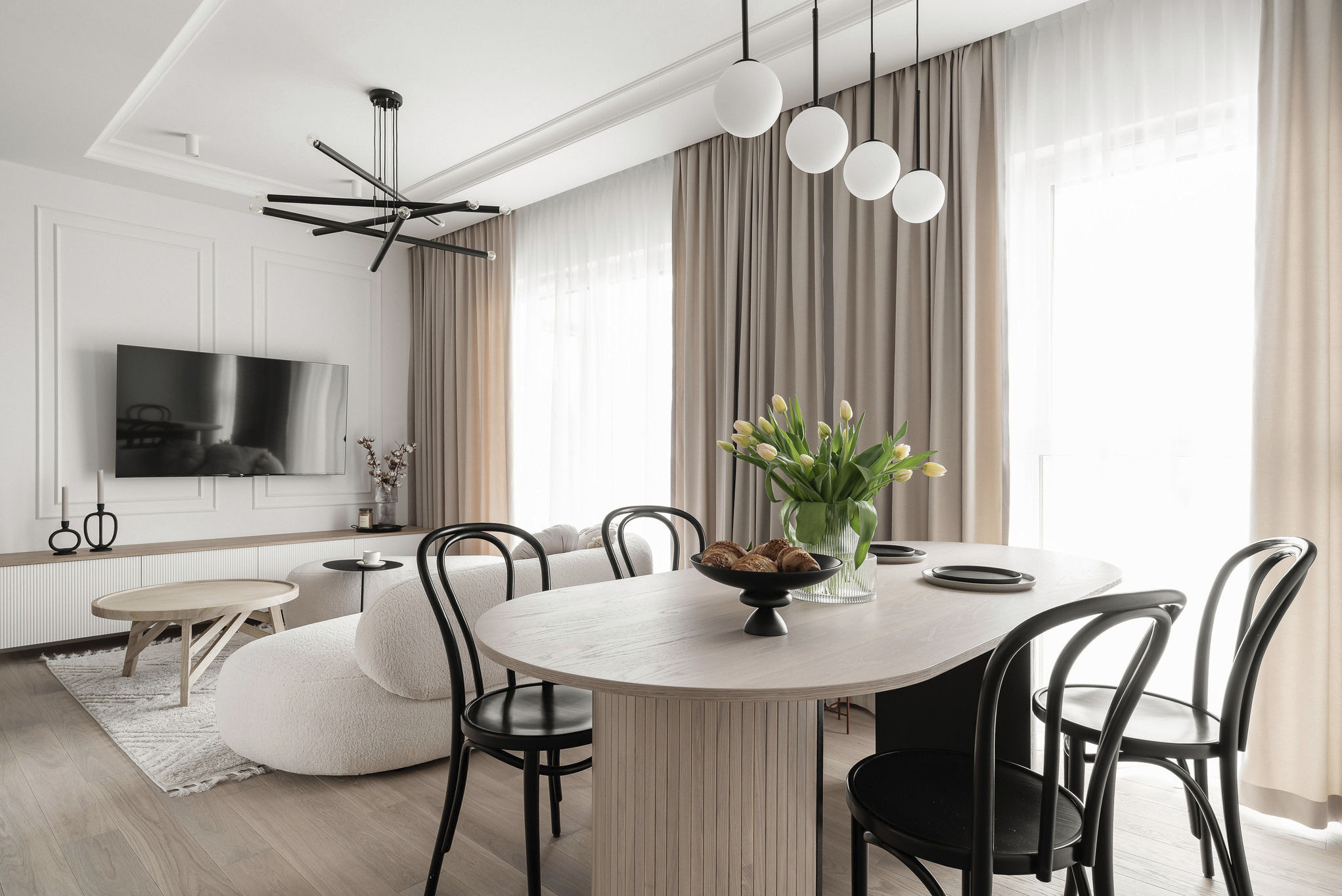 Jasne wnętrze z owalnym stołem, czarnymi krzesłami i bukietem tulipanów. W tle sofa boucle i nowoczesna lampa sufitowa.