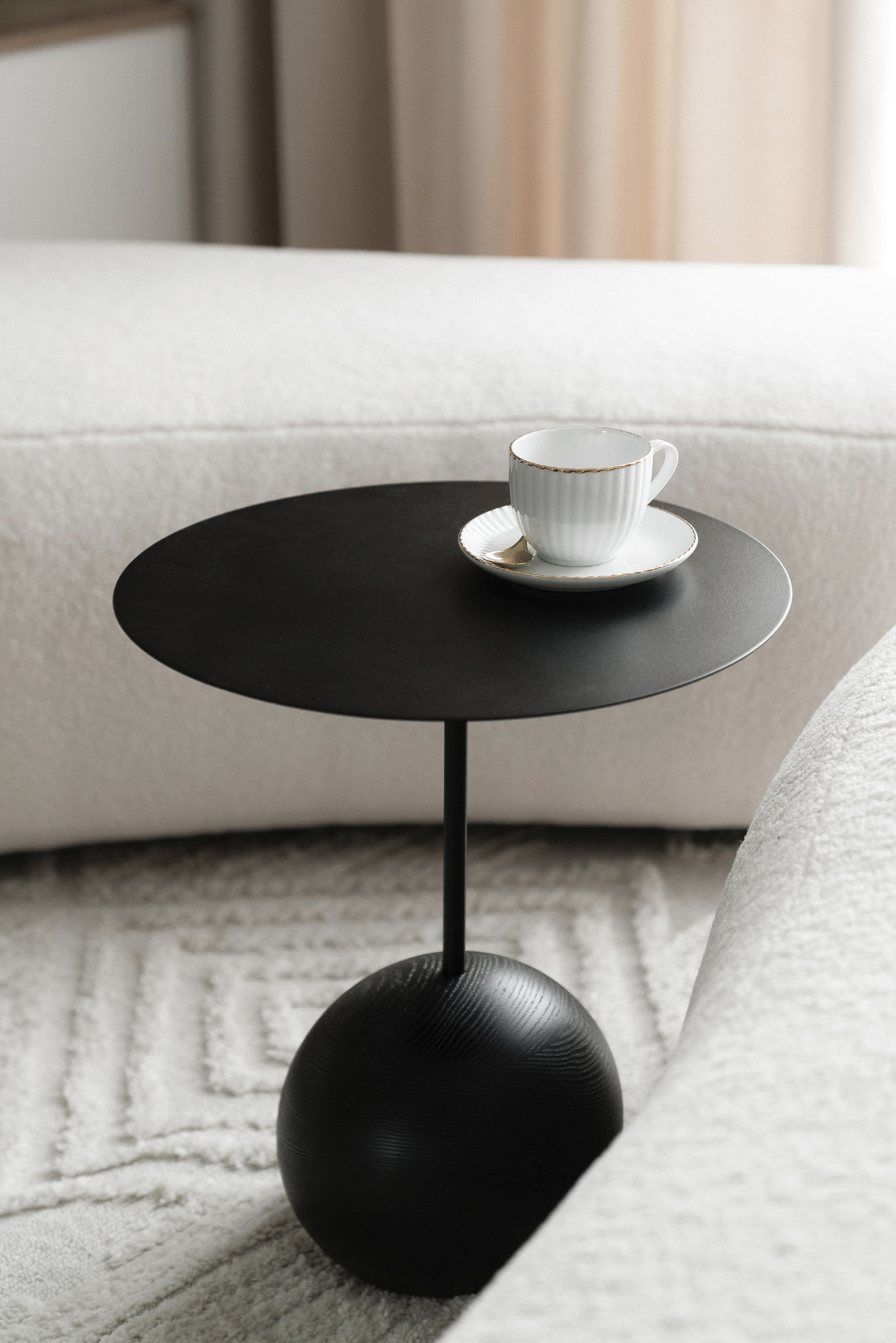 Minimalistyczny stolik kawowy z czarnym blatem i kulistą podstawą, na którym stoi filiżanka z sosem, na tle kremowej sofy i dywanu.