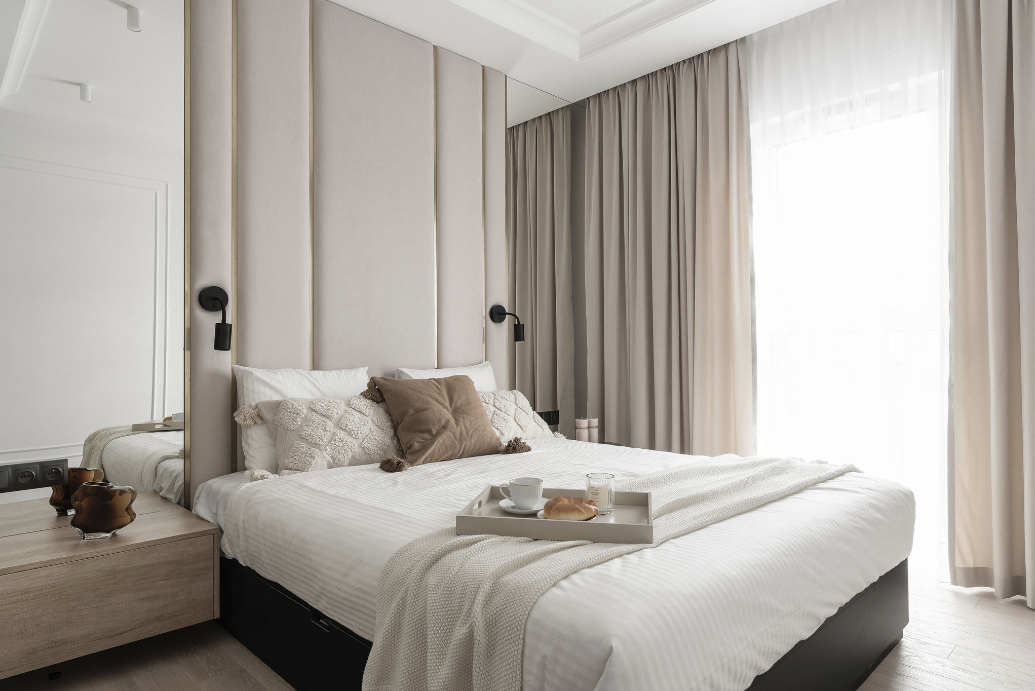 Elegancka sypialnia w jasnych barwach z tapicerowanym wezgłowiem i śniadaniem na łóżku. Minimalistyczne lampki nocne, lustro i subtelne zasłony.