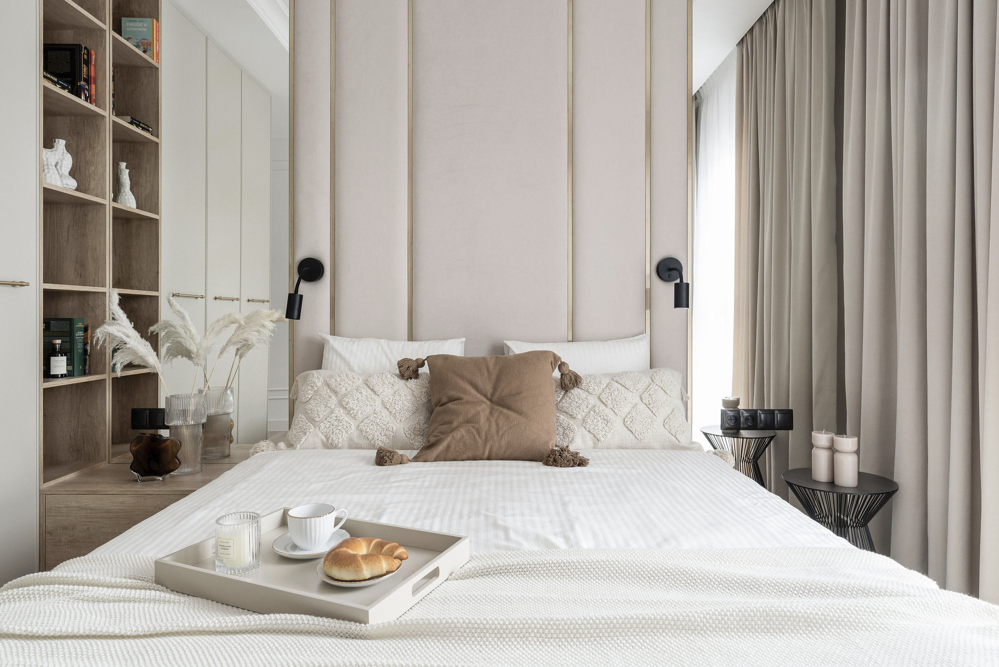 Jasna sypialnia z beżowym zagłówkiem, dekoracyjnymi poduszkami i śniadaniem na tacy. Minimalistyczny styl z akcentami w odcieniach beżu i bieli.