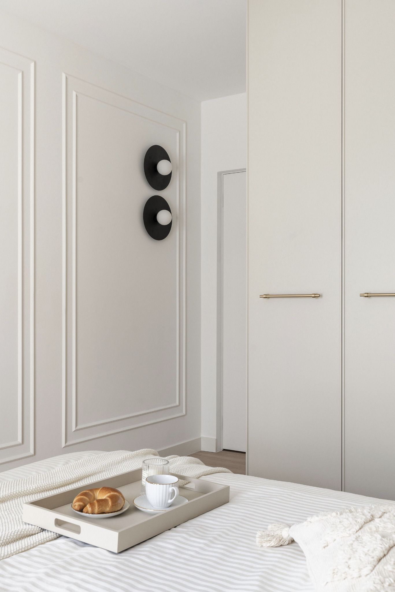Jasna sypialnia: łóżko z śniadaniem na tacy, minimalistyczne lampy ścienne i biała szafa z złotymi uchwytami. Klasyczne sztukaterie na ścianach, spokojna kolorystyka.
