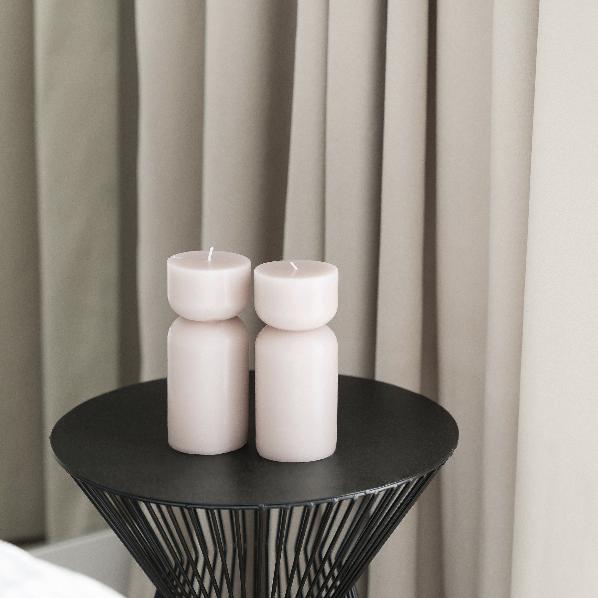 Dwie geometryczne, różowe świece na czarnym, drucianym stoliku, na tle beżowych zasłon. Minimalistyczny detal wnętrza.