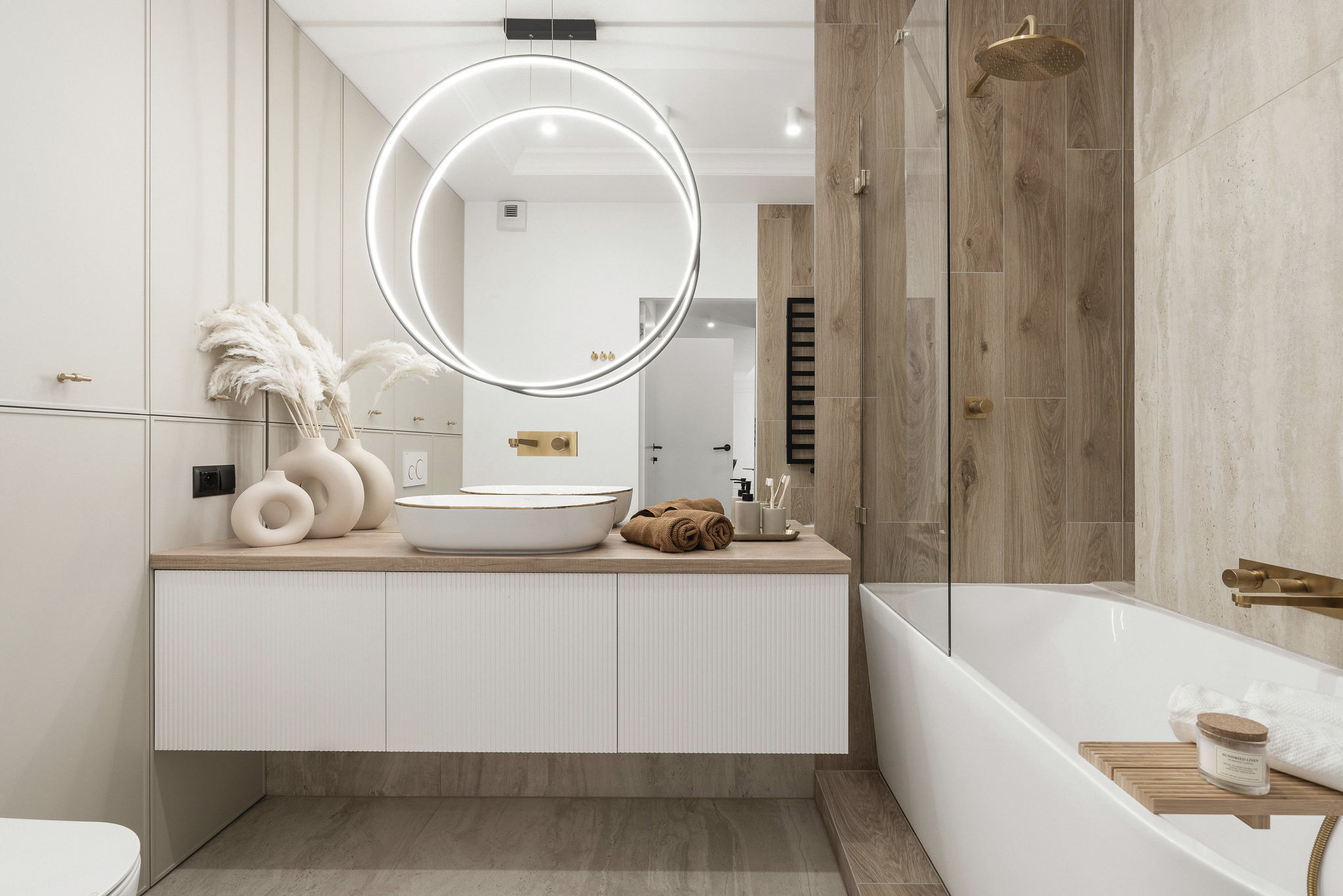 Stylowa łazienka z drewnianymi akcentami: okrągłe lustro z podświetleniem LED, biała umywalka nablatowa i armatura w kolorze złota.