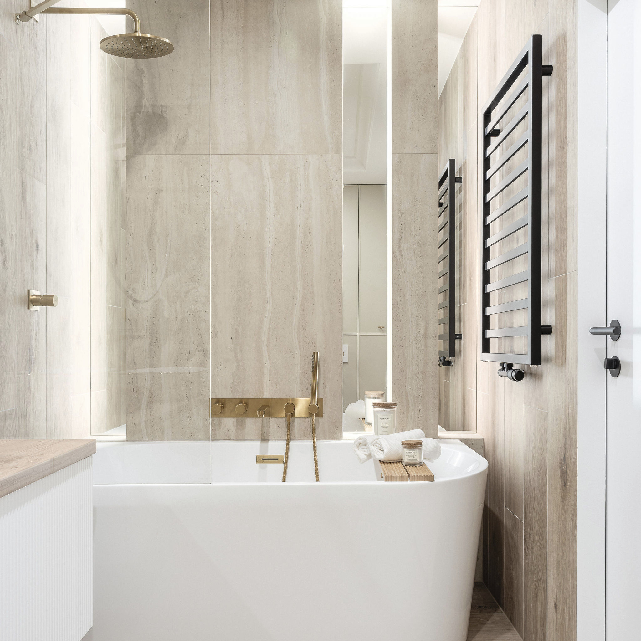 Elegancka łazienka w beżach z wolnostojącą wanną, złotą armaturą i czarnym grzejnikiem drabinkowym, oświetlona listwami LED.