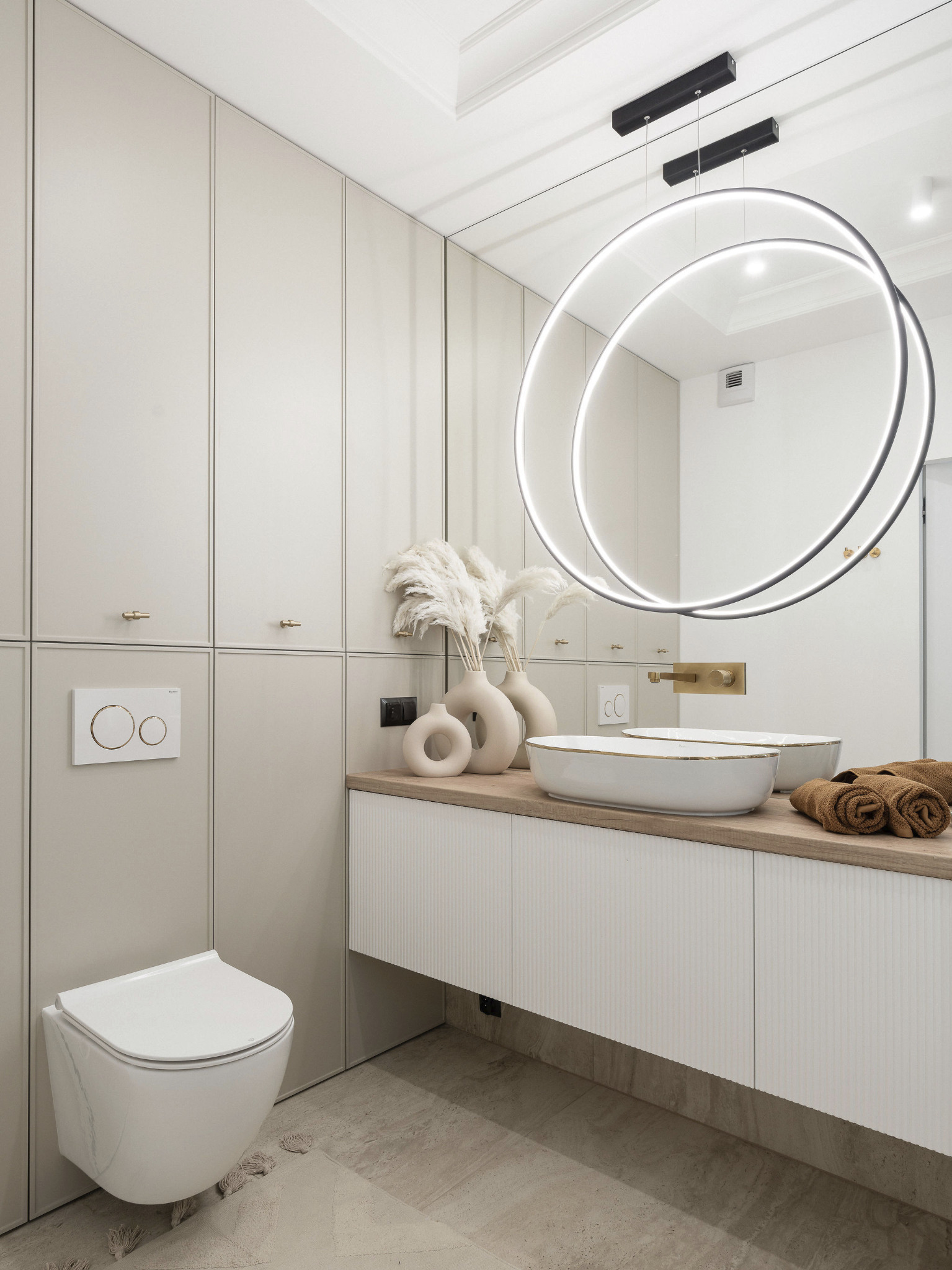 Elegancka łazienka w beżach: wisząca toaleta, umywalki nablatowe z dekoracyjnym oświetleniem i szafki w zabudowie, tworzące spójną i funkcjonalną przestrzeń.