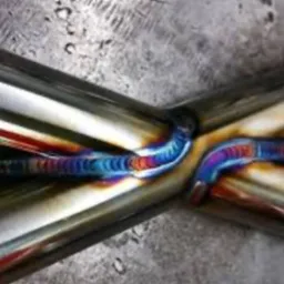 Precyzyjne spawanie TIG rur nierdzewnych, widoczne tęczowe przebarwienia spoiny, świadczące o wysokiej jakości wykonania. Szlifowana powierzchnia metalu w tle.