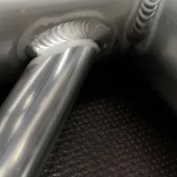 Szczegółowe zbliżenie na precyzyjne spawy łączące metalowe rury. Widoczne regularne ściegi spawalnicze, wysoka jakość wykonania. Metal o srebrzystym odcieniu.