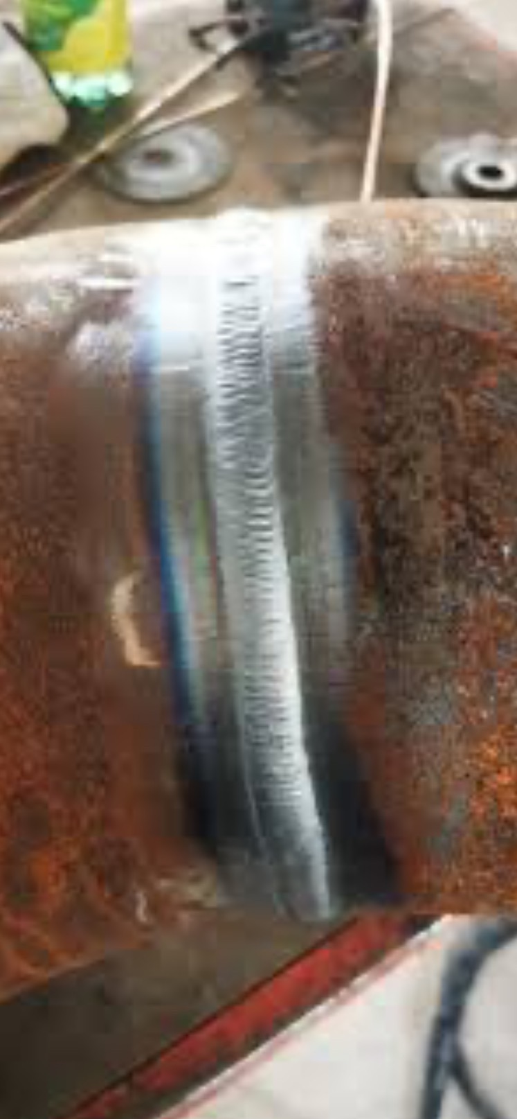 Spoina na rurze z widocznymi śladami ciepła i przebarwieniami. Tło stanowią narzędzia i akcesoria warsztatowe, sugerujące proces obróbki metalu.