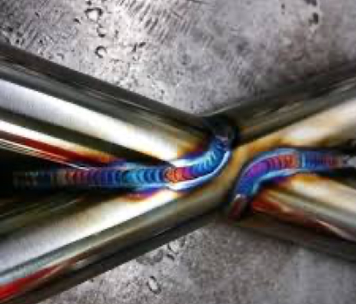 Precyzyjne spawanie TIG rur nierdzewnych, widoczne tęczowe przebarwienia spoiny, świadczące o wysokiej jakości wykonania. Szlifowana powierzchnia metalu w tle.