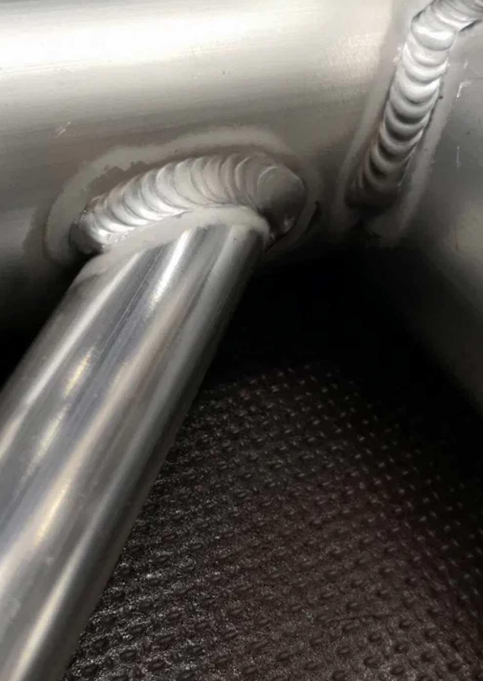 Szczegółowe zbliżenie na precyzyjne spawy łączące metalowe rury. Widoczne regularne ściegi spawalnicze, wysoka jakość wykonania. Metal o srebrzystym odcieniu.