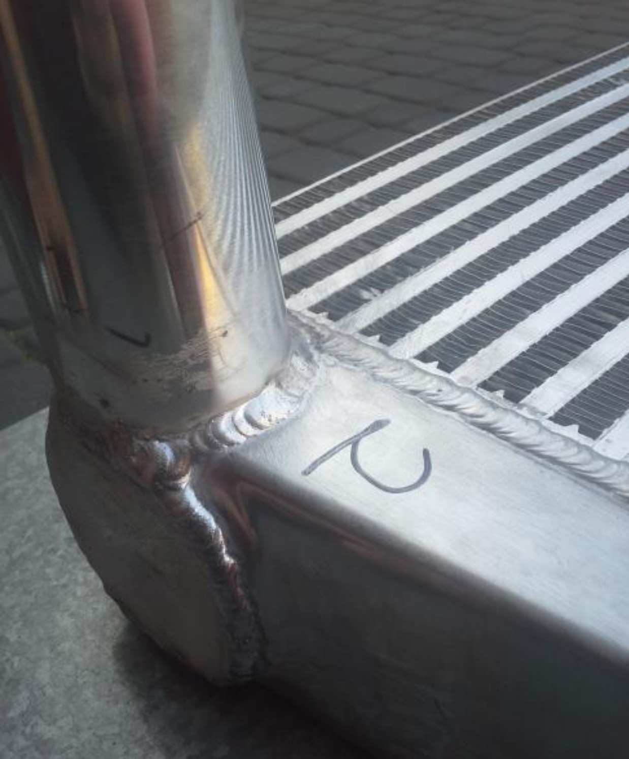 Szczegółowe ujęcie spawu łączącego elementy metalowe, z widocznymi śladami obróbki i oznaczeniami. Widoczna faktura metalu i precyzyjne wykonanie.