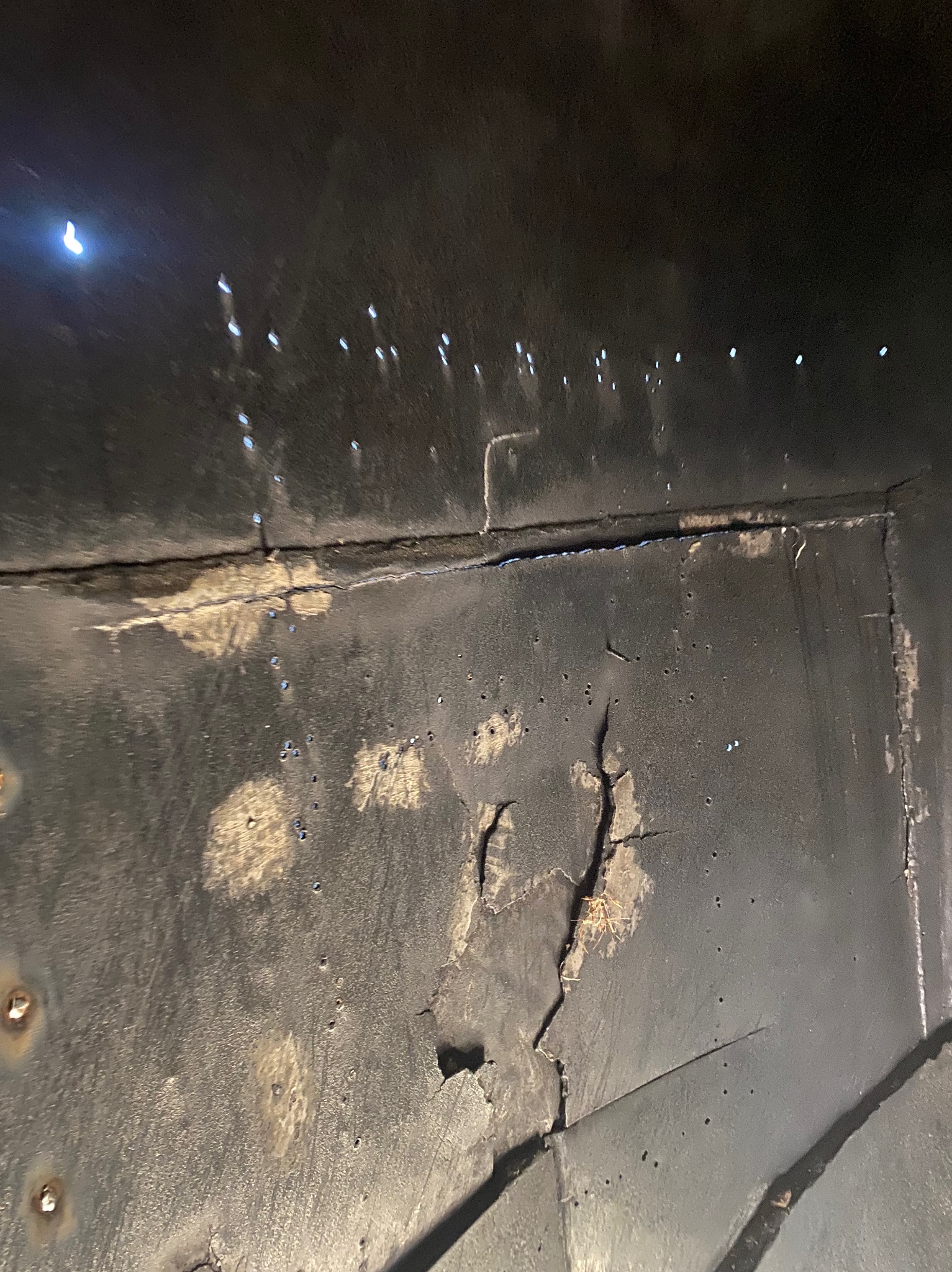 Fragment czarnej, metalowej konstrukcji z widocznymi śladami korozji i pęknięciami, oświetlony punktowo. Widoczne nity i spawane łączenia.