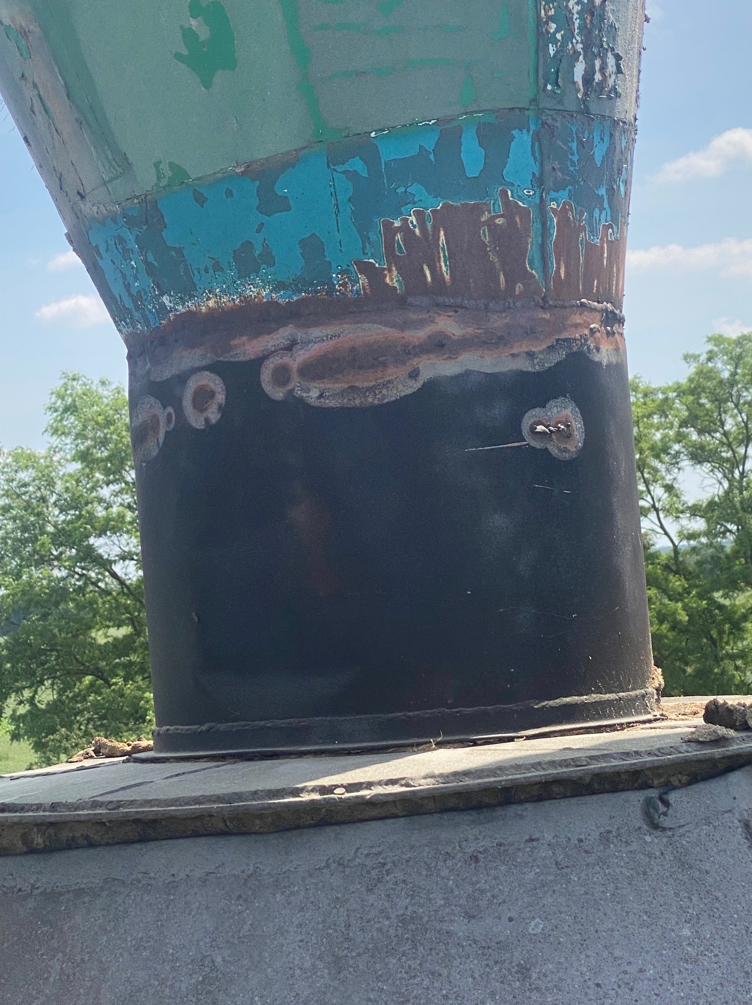 Cylindryczna konstrukcja metalowa z widocznymi śladami korozji i odpryskującą farbą, z detalami spawów i nitów. Tło stanowią drzewa i niebo.