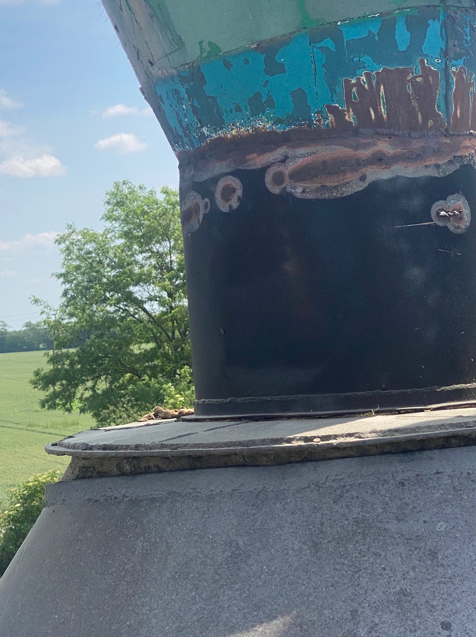 Fragment metalowej konstrukcji z widocznymi śladami spawania, rdzy i łuszczącej się farby, na tle zieleni i błękitnego nieba. Detal z bliska, ukazujący strukturę i niedoskonałości materiału.